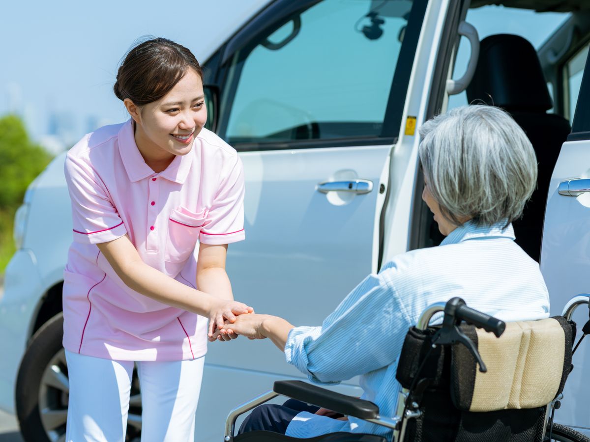 福祉車両の横で、車いすの高齢の女性の手を取るヘルパー