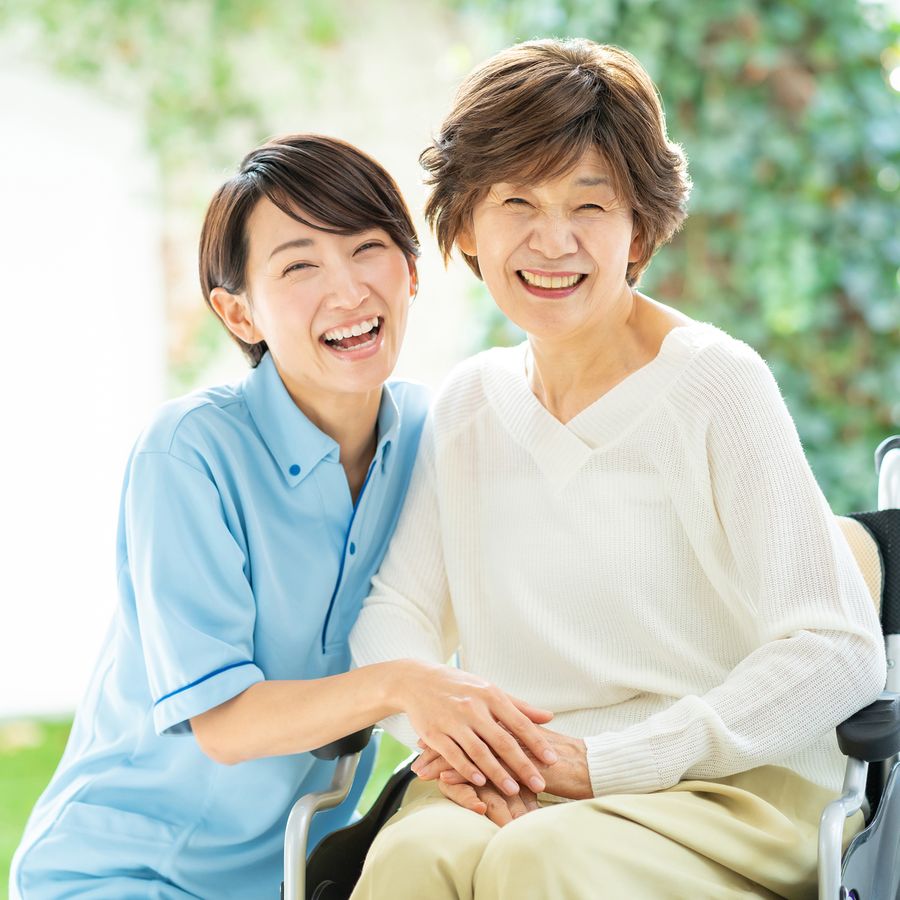 庭先で、車いすに座る高齢の女性と肩を寄せて笑うヘルパー