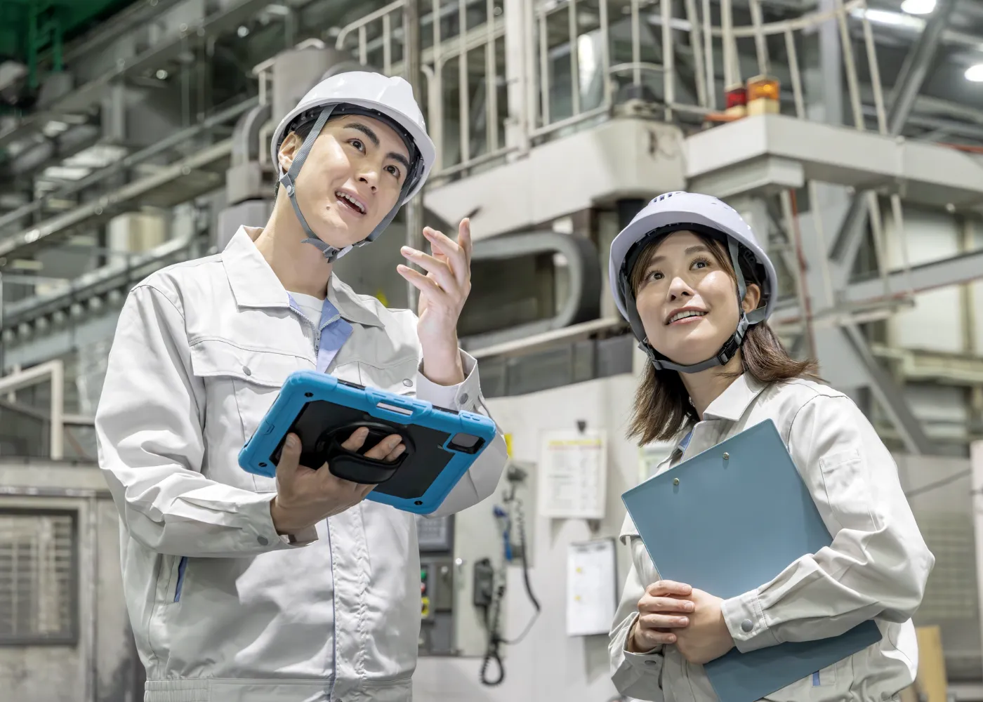 ヘルメット姿の先輩社員が、タブレットを手に工場の設備を指さしながら後輩に説明している